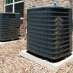 Two air conditioning units on concrete bases, surrounded by mulch and a brick wall, from Chad's AC Direct.