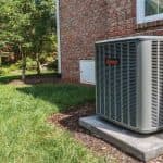 Air conditioning unit on a concrete pad, surrounded by grass and trees, against a brick house exterior.
