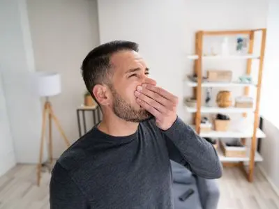 Man covering his mouth and nose, appearing uncomfortable due to poor indoor air quality in a modern living room, emphasizing the need for air duct cleaning services.