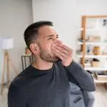Man yawning in a modern living room with a tripod lamp and decorative shelf, showcasing indoor comfort at Chad's AC Direct.
