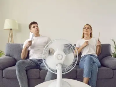 Man and woman sitting on a couch fanning themselves with handheld fans, facing a table fan, illustrating discomfort with indoor heat and the need for effective air conditioning solutions.