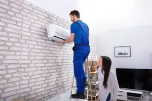 HVAC technician in blue uniform installing air conditioning unit on wall while homeowner observes, highlighting professional service for efficient furnace and HVAC system repairs.