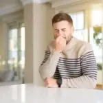 Young man in striped sweater sits at a white table in a minimalist room, highlighting HVAC service discussions for summer.