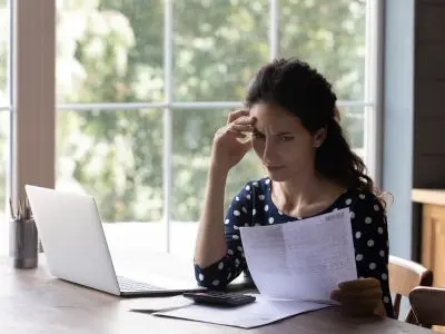 Woman with a concerned expression reviewing utility bills and paperwork at a table, highlighting the impact of high heating costs due to an inefficient heating system.