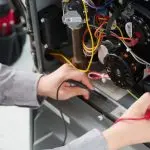 Technician working on HVAC machinery with multi-meter and colorful wires in a workshop setting.