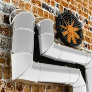 Air duct system with metal piping and ventilation fan against a brick wall, illustrating HVAC components for efficient air delivery in homes.