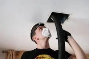 Technician using a vacuum hose to clean a ceiling air duct, emphasizing HVAC maintenance and air quality improvement.
