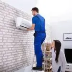 Technician repairing air conditioning unit at a residential property, overseen by an attentive customer for Chad's AC Direct.