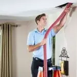 Man in a blue shirt using a ladder for HVAC maintenance, pulling a red hose from the ceiling for repairs.