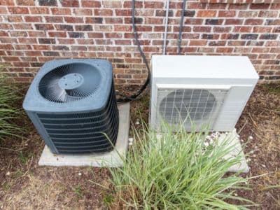 Air conditioning units outside a home, featuring a central air conditioner on the left and a ductless mini-split unit on the right, surrounded by grass and a brick wall.