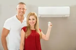 Couple standing next to a wall-mounted central air conditioning unit, woman holding a remote control, smiling, illustrating the benefits of modern HVAC systems and comfort in home cooling.