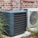 Air conditioning units outside a home, featuring a dark gray unit and a white unit near greenery and a brick wall.