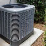 Air conditioning unit on concrete slab with gray exterior and ventilation lattice, surrounded by green foliage.