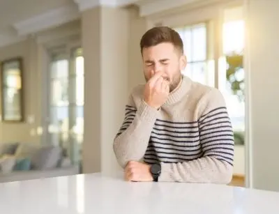 Man sitting indoors, grimacing and pinching his nose, indicating discomfort from musty odors in a humid home environment.