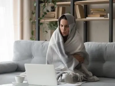 Woman wrapped in a blanket, looking uncomfortable while sitting on a couch with a laptop, illustrating the discomfort of a poorly heated home.