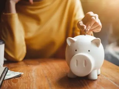 Piggy bank on wooden table with person inserting coin, symbolizing savings and financial planning.