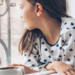 Young girl in casual shirt gazes out window with coffee cup, showcasing a tranquil moment.