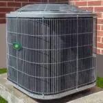 Air conditioning unit on a concrete slab, featuring a grey metallic exterior and grilled enclosure, near a brick wall.