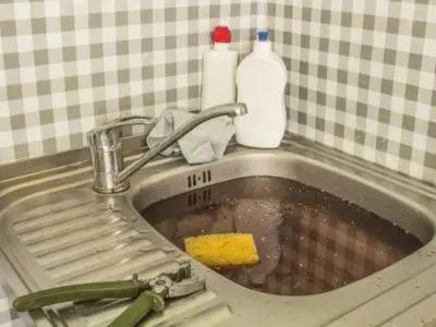 Clogged kitchen sink filled with dirty water, sponge, and cleaning supplies, indicating the need for professional drain cleaning services.