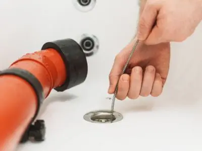 Hand using a plumber's snake to clear a drain, with a red drain pipe visible, illustrating professional drain cleaning techniques.