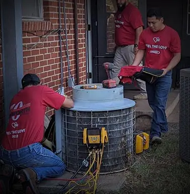 Technicians servicing an air conditioning unit, wearing red "Feel the Love" shirts, demonstrating HVAC maintenance expertise in a residential setting.
