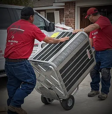 Two technicians in red shirts transporting a large air conditioning unit on a cart, emphasizing HVAC installation and service in the Midland-Odessa area.