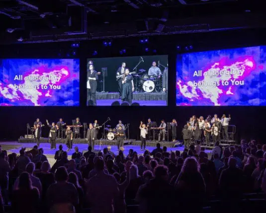 Worship service at Odessa Christian Faith Center with a choir and band performing, large screens displaying lyrics "All of the glory belongs to You," and an engaged audience.