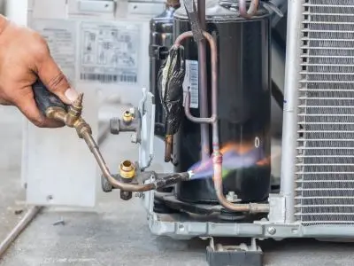Technician performing maintenance on HVAC unit, using a torch on refrigerant lines, illustrating the need for frequent repairs in older systems.