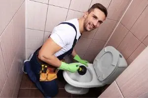 Man in green gloves cleaning a toilet with a plunger, emphasizing plumbing maintenance and the importance of only flushing toilet paper.