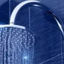 Chrome showerhead with water droplets on a blue tiled wall, by The Meridian Company.