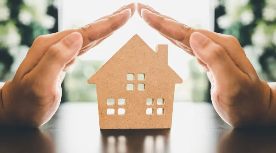 Human hands protecting a crafted house model on a table, representing home security and maintenance by The Meridian Company.