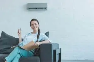 Woman sitting on a couch holding a remote control, smiling, with a ductless HVAC mini-split system mounted on the wall in a modern living room setting.