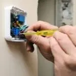 Close-up of hands using a screwdriver on a thermostat for HVAC electrical work, highlighting organized wiring and tools.