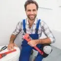 Smiling technician in blue overalls holds a red pipe wrench in a bright bathroom, representing The Meridian Company.