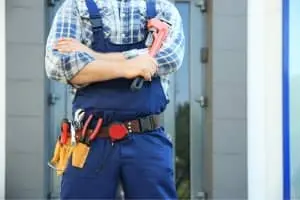 Plumber in blue overalls holding a wrench, equipped with various tools, standing in front of a modern building, representing plumbing services offered by The Meridian Company.