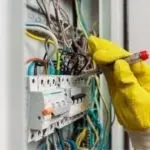 Human hand in a yellow glove repairing an electrical panel with colorful wiring and a screwdriver for HVAC maintenance.