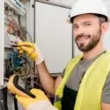 Smiling male electrician in hard hat and safety vest, using multimeter on electrical wiring in a service area.