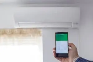Person using smartphone to control a smart air conditioning unit on the wall, illustrating modern HVAC technology and home cooling solutions.