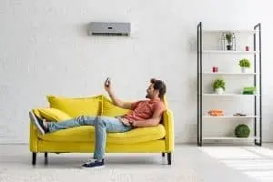 Man relaxing on a yellow couch using a remote control, with a ductless HVAC system mounted on the wall, illustrating comfort and climate control in a modern living space.