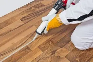 Person applying caulk to seal gaps in wooden flooring, emphasizing HVAC maintenance and air leak prevention.