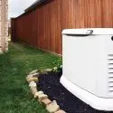 White HVAC unit surrounded by landscaped stones and mulch near grass and a wooden fence, from The Meridian Company.