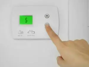 Thermostat display with a green screen showing a dollar sign, hand adjusting settings, relevant to HVAC maintenance and energy efficiency tips.