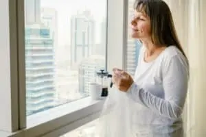 Woman inspecting window for insulation improvements, holding window insulation film, overlooking a cityscape, emphasizing energy efficiency and home comfort.