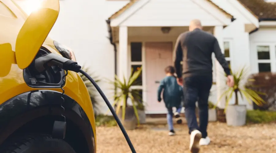 Electric vehicle charging station connected to a yellow car with a family approaching their home, highlighting the convenience of home EV charging solutions.