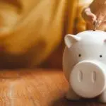 White piggy bank with a hand inserting a coin on wood, showing savings for home maintenance by The Meridian Company.