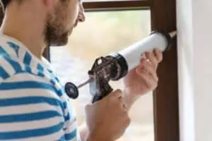 Man applying weather-resistant caulk to window frame for energy efficiency and heat retention.