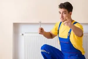HVAC technician in blue overalls holding a wrench, pointing at the viewer, with a radiator in the background, representing HVAC maintenance and air quality solutions by The Meridian Company.