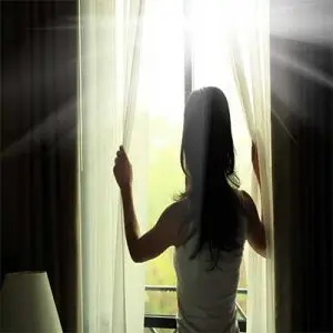 Woman opening sheer curtains to let in natural light, symbolizing home comfort and improvement.