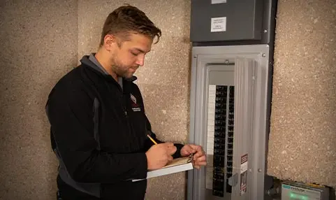 Electrician inspecting a circuit breaker panel and taking notes, representing The Meridian Company's electrical services.