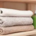 Rolled towels in white, gray, and beige on a wooden shelf, with a green bottle, promoting cleanliness for homeowners.